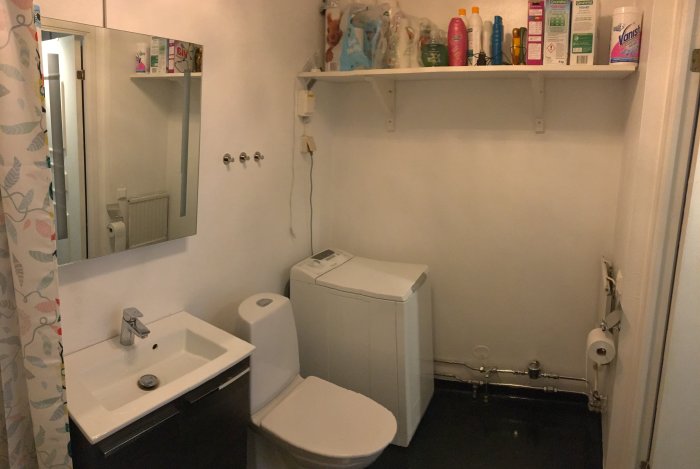 Äldre badrum med vita väggar, spegelskåp, handfat, toalett och toppmatad tvättmaskin, inför renovering.
