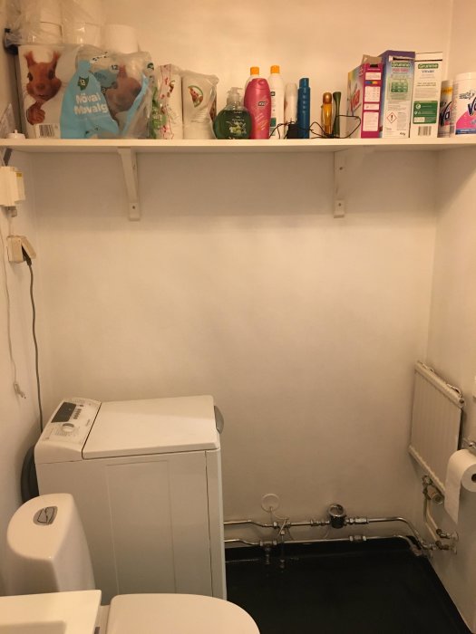 Bild på ett badrum före renovering med tvättmaskin, toalett, hylla med badrumsprodukter, och synliga rör på väggen.