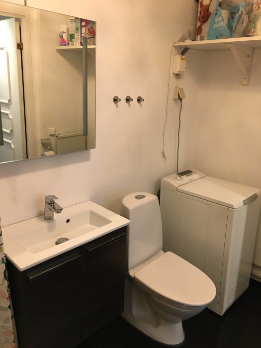 Badrum i behov av renovering med vägghängt skåp, handfat, toalett och tvättmaskin.