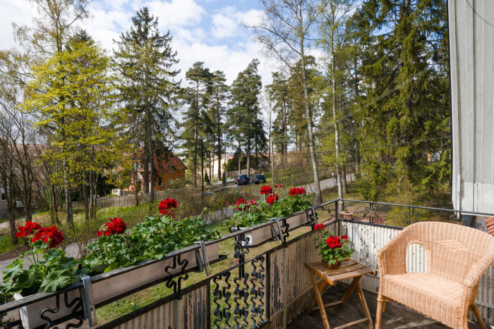 Utsikt från en balkong med röda blommor och rottingmöbel, omgiven av grönska och villa i bakgrunden.