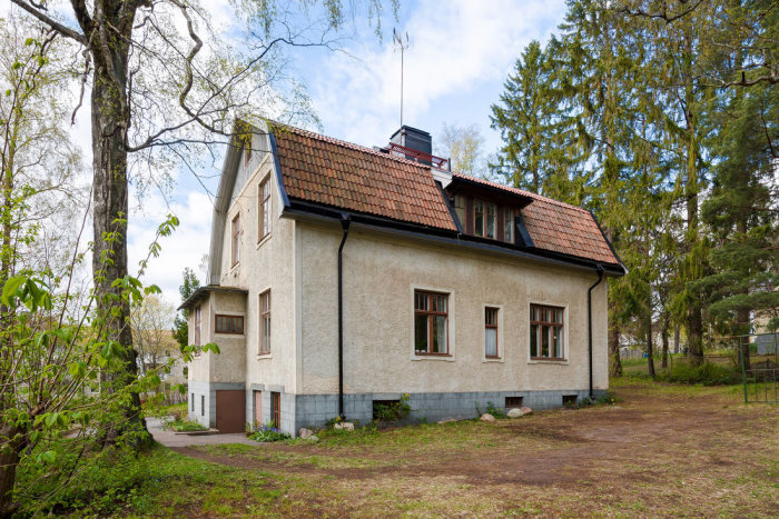 Tvåvåningshus med putsad fasad, röd tegeltak och omgivande trädgård i dagsljus.