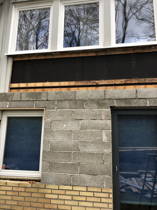 Delvis putsad leca-blockvägg och oputsad del med asfaltboard under fönster på en husfasad.