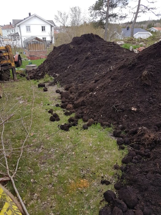 Stor hög med matjord på en gräsmatta med hus och växthus i bakgrunden.