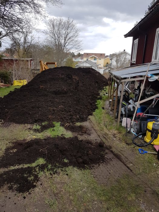 En stor hög med matjord bredvid en grävare i en trädgård, redo för landskapsarbete.