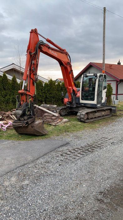 Grävmaskin från Kubota parkerad i en trädgård, omgiven av grus, tegelstenar och en barnlekcykel.