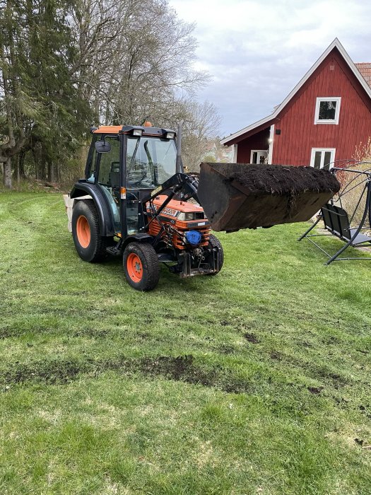 Traktor med frontlastare full av matjord på en gräsmatta med röd stuga i bakgrunden.