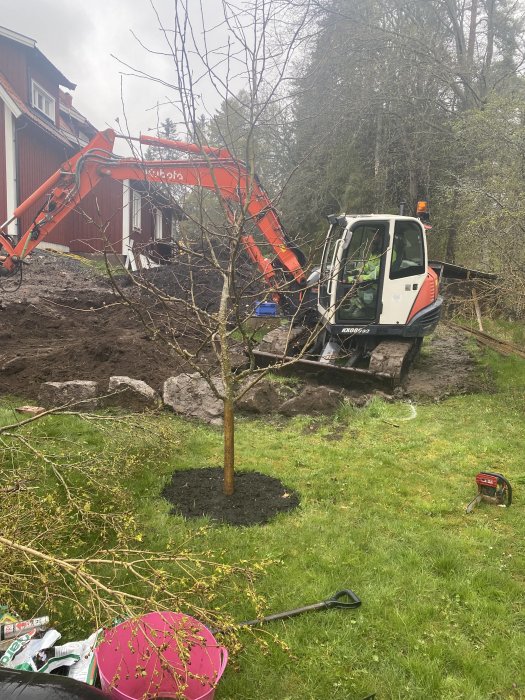 Orange grävmaskin av märket Kubota i arbete i en trädgård, med stor hög jord och avtäckt berg.