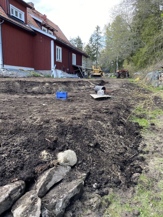 Nyutlagd matjord i trädgård med grävmaskin i bakgrunden, berg och planerade odlingslådor nära röd stuga.