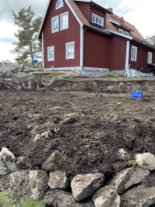 Rödfärgat hus med avtäckt berg och nylagd matjord på trädgårdstomt, förberett för poolbygge.