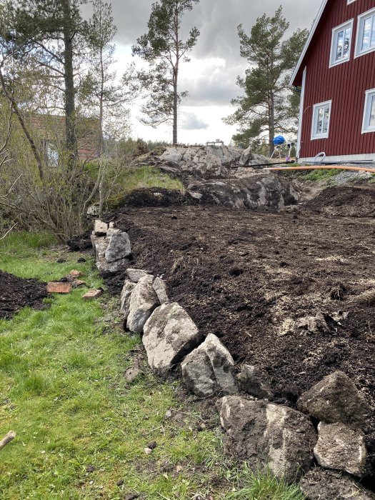 Nyjordad trädgårdsterrass med avtäckt berg och stenrad nära rött hus.