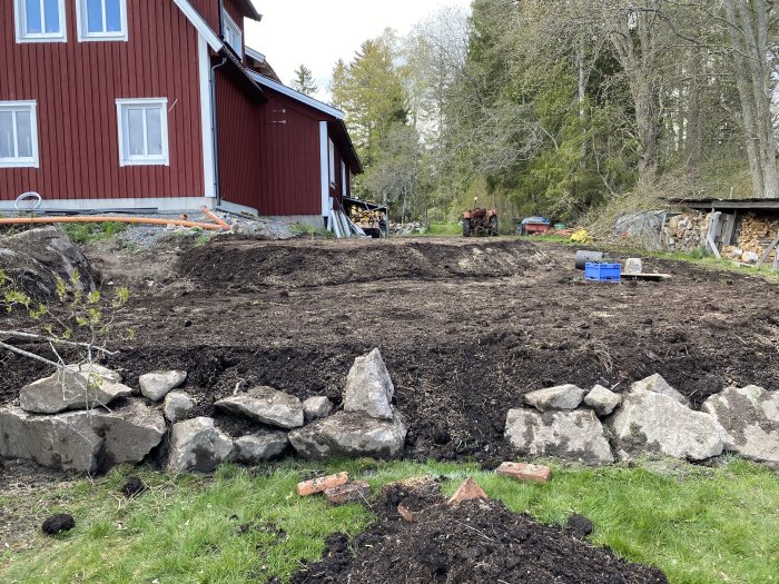 Nyplanterad matjord i två nivåer med avtäckt berg nära röd husgavel, stora stenar i förgrunden.