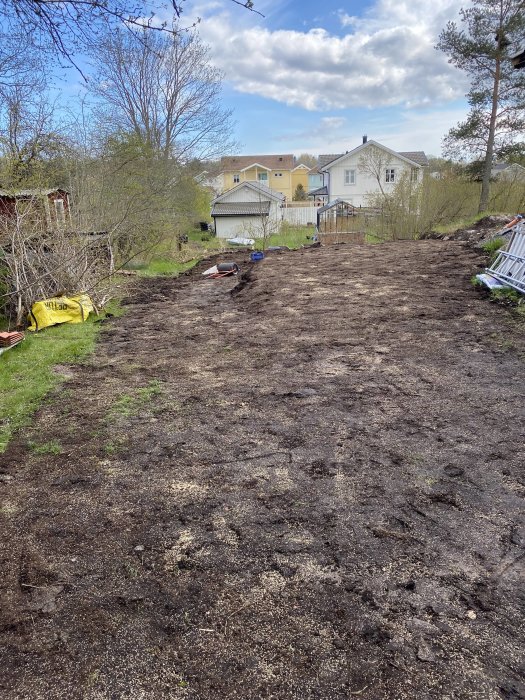 Nyplanterat område med matjord och avtäckt berg, arbetsredskap syns och hus i bakgrunden.