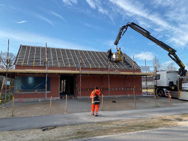 Byggarbetsplats med arbetare, byggnadsställningar och en kranbil som levererar takstolar till ett hus under konstruktion.