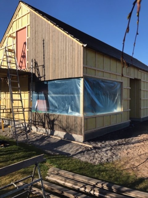 Delvis uppbyggt trähus med isolering, byggställning och täckt med en presenning under en klar himmel.