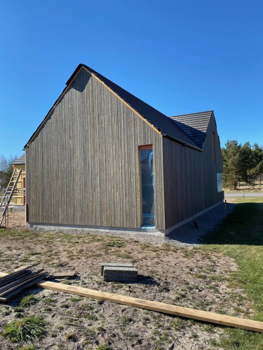 En nybyggd trähusgavel med stående panel, takavvattning och grävda tomtytor framför.