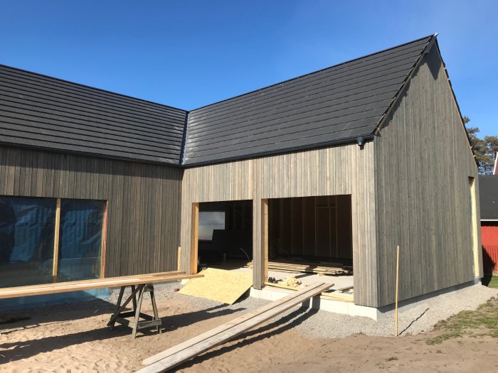 Nybyggt hus med träpanel, svart tak och pågående byggnadsarbete, byggmaterial på marken.