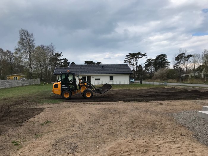 Minigrävare som planerar ut jord på en byggplats med nybyggd vit hus i bakgrunden.