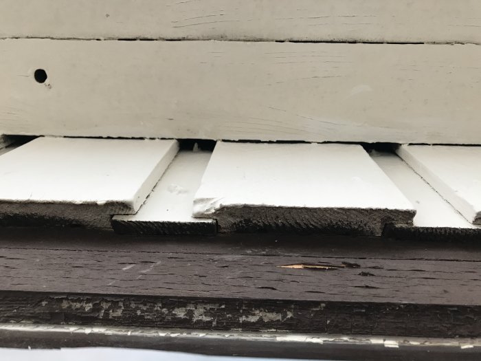 Närbild på slitet träpanel med vitmålad fasskråpanel på en byggnad.