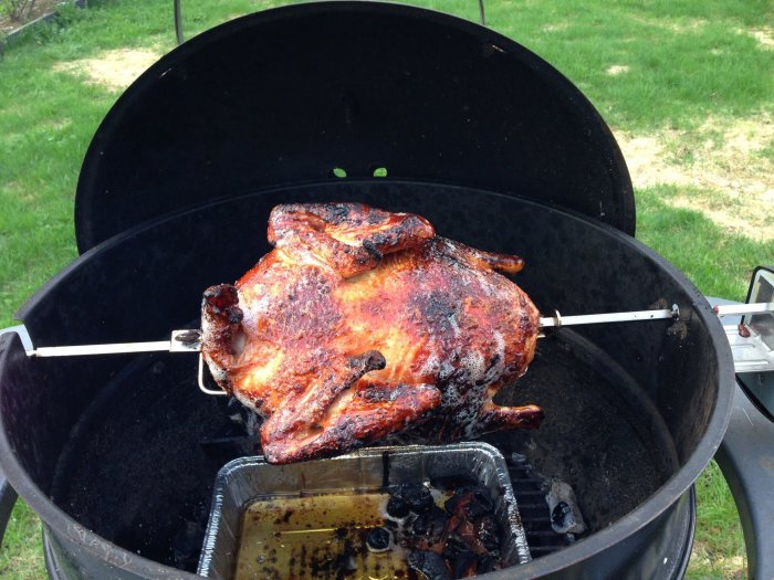 Grillad kyckling på spett i en öppen Weber-grill med droppskydd och kol under.