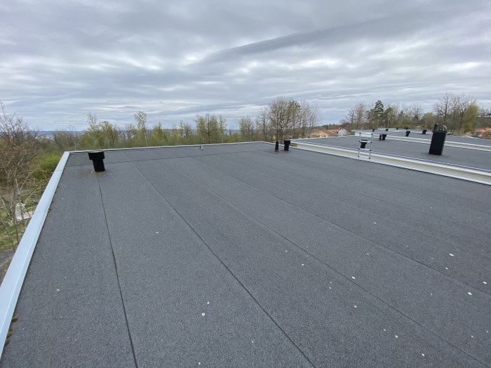 Platt tak med svart takpapp, ventilationsrör och skyddsräcken mot en molnig himmel.