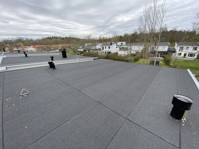 Flacktak med mörka takplattor, ventilationsrör, solpaneler och utsikt över bostadsområde.