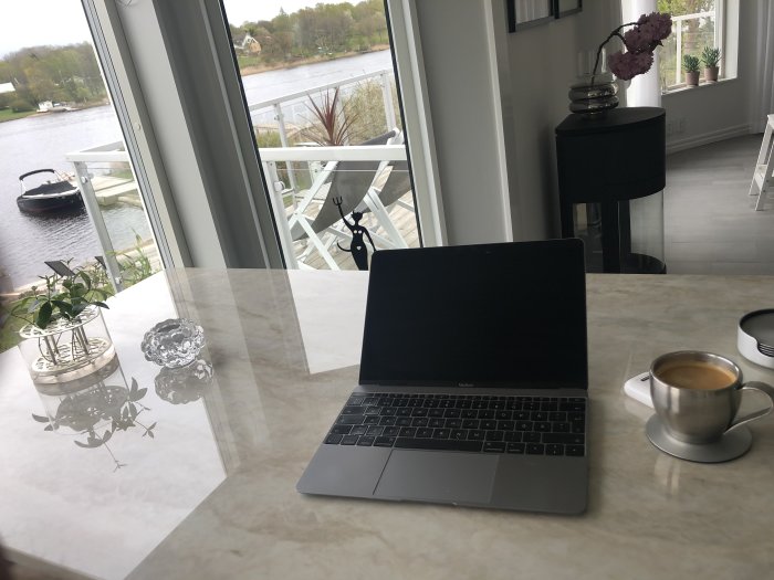 Arbetsplats hemma med öppen laptop, kaffekopp och utsikt över vatten och båt från ett fönster.