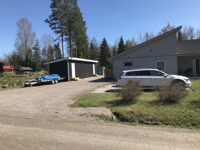 Infart med grus, parkerad bil och släpvagn framför en bostad och garage.