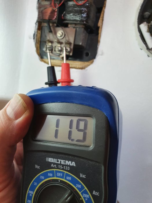 Multimeter som visar 11,9 volt med en demonterad transformator i bakgrunden.