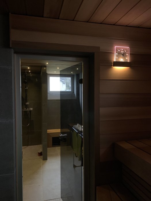 Utsikt från en bastu med tända LED-lyktor och temperaturmätare, alulister runt dörren och inblick i duschrum.