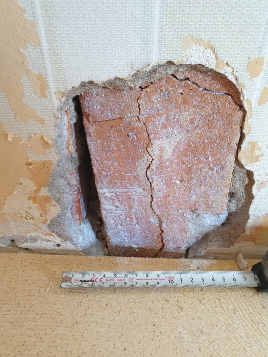 Ett hål i en murstock med lossad puts och synliga tegelstenar, med ett måttband framför för storleksreferens.