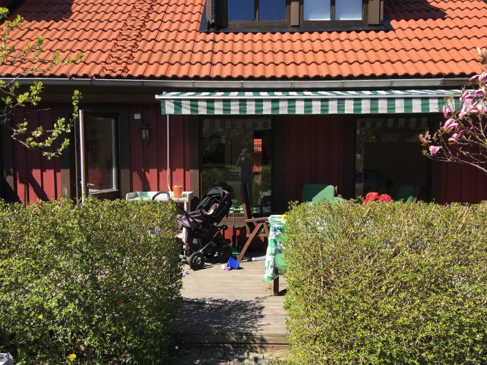 Tradäck framför ett rött radhus med tegeltak, möbler och en barnvagn, omgivet av grönska.