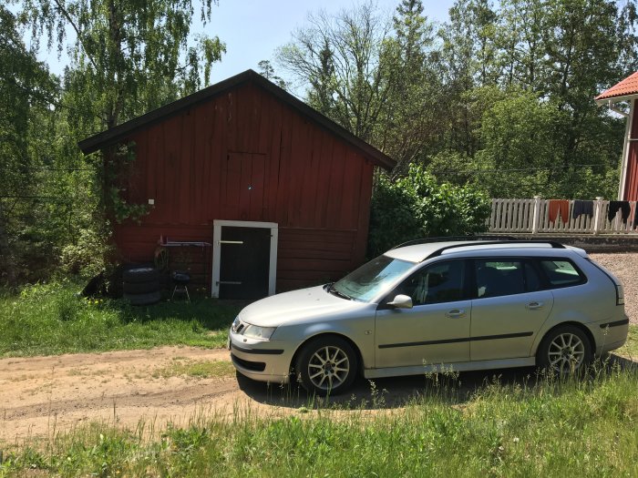 Gammal röd bod med plåttak och en vit dörr, nära en silverfärgad bil och grönska.