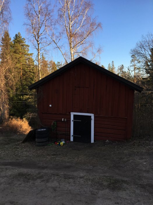 Gammal röd tvåvåningsbod med modern plåttak, slitet utseende och öppen dörr, omgiven av träd och barnleksaker framför.