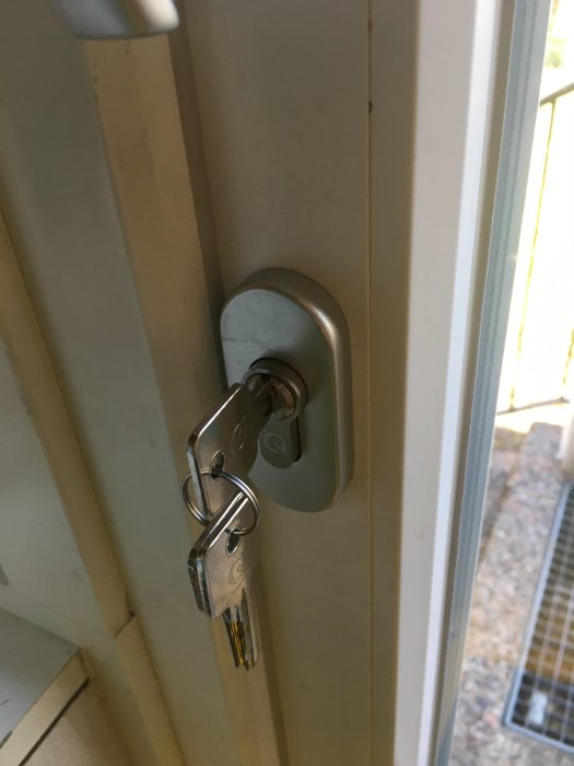 Nycklar i låscylinder på en vit dörr utan synliga skruvar.