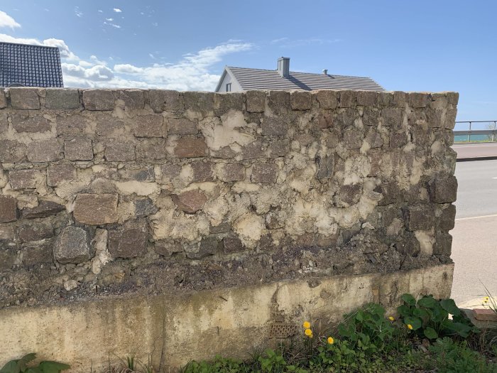 Mur av sten och bruk med ojämnt fundament synligt nederst, omgivet av grönska.