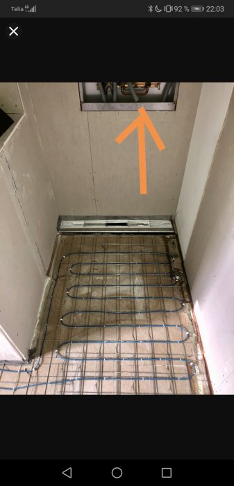 En ofärdig dusch med öppen väggpanel som visar en installationslåda, pekad på av en orange pil, och golvvärmeledningar.