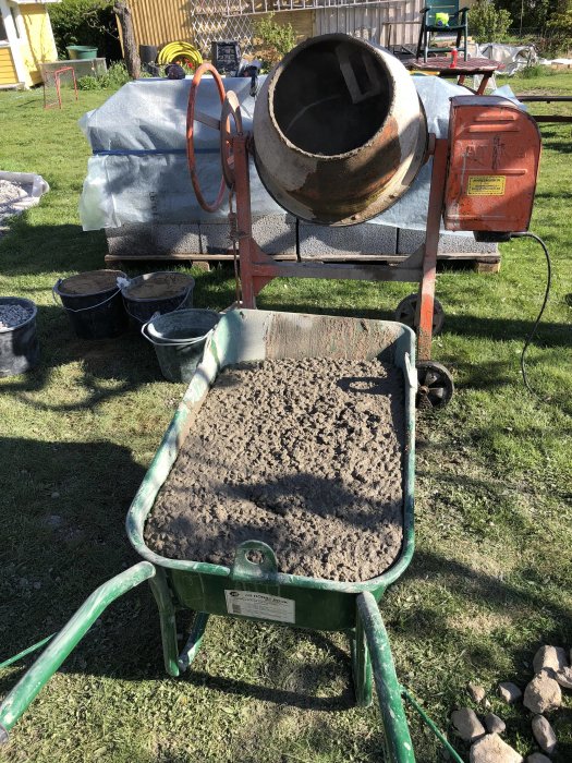 En blandare för cement och en hjulbarrow fylld med färsk betong i ett trädgårdsbyggprojekt.