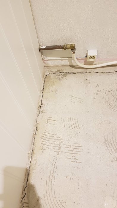 Spricka i betonggolv intill kant med vägg, golvvärmerör och strömuttag syns.