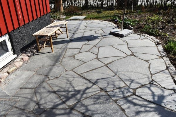 Oregelbunden offerdalskiffer som stenläggning på en trädgårdsgång med bänk och husvägg i bakgrunden.