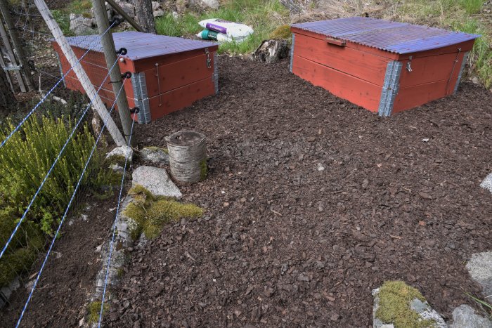 Trädgårdsyta med täckbark och två röda kompostbehållare bakom blått rådjursnät och gröna buskar.