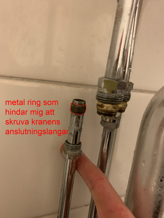 Läckande koppling mellan blå och röd vattenledning under handfatet markerad av ett finger, behöver tätning.