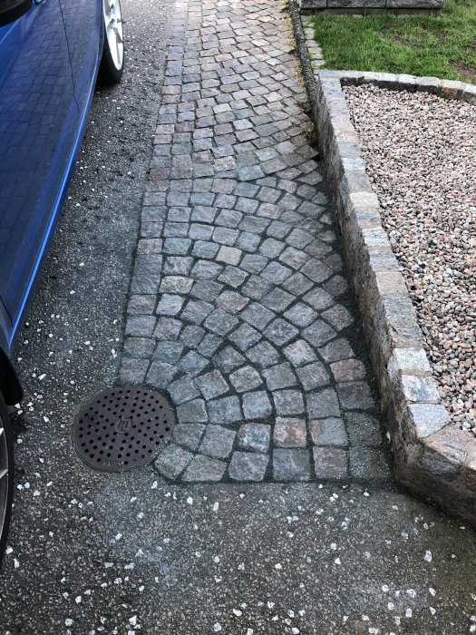 Rensade fogar mellan gatsten vid en uppfart nära en blå bil och sidokant i sten med grus.
