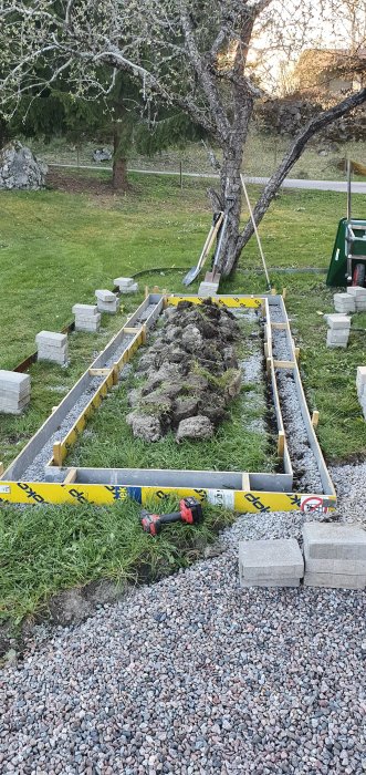 Pågående bygge av gjutform för odlingslåda med spadar, grus och betongblock i en trädgård.