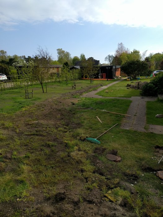 Trädgård under anläggning, med igenväxande grävda fåror och en räfsa på marken, bakgrunden visar ett hus och träd.