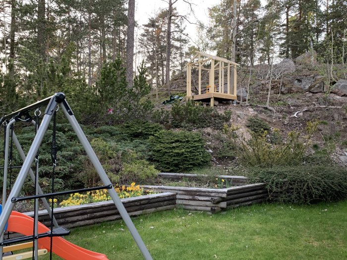 Bild på en trädgård med gungställning i förgrunden och en lekstuga ovanpå en bergsknalle i bakgrunden.