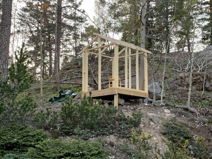 Ofärdig trälekstuga på pelare i skogsmiljö med murknande slipers och stenar på marken som kan användas för trappbygge.