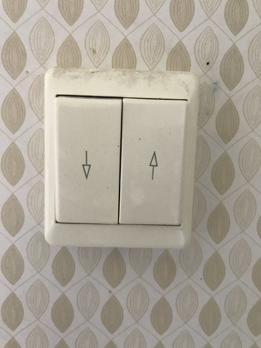 Väggströmbrytare med upp och ner pil symboler på framsidan, smutsig och använd, monterad på tapetserad vägg.