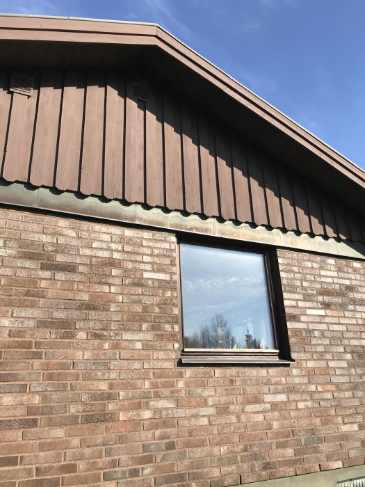 Del av husfasad med mörkbruna paneler och tegelvägg under blå himmel.