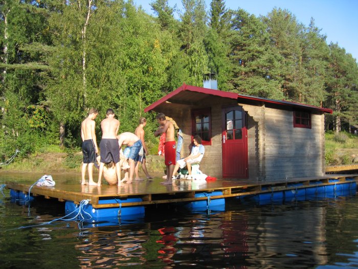 Bastuflotte med personer på en sjö vid en skogsbryn, stabil konstruktion utan motor.