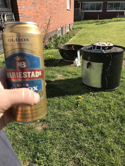 En hand håller en ölburk med Mariestads Old Ox öl framför en trädgård där en grill med kyckling på står.